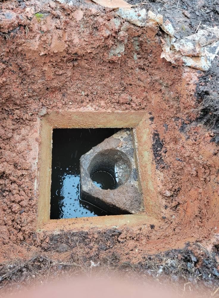 Square concrete utility opening in muddy ground with a pipe and pooled water inside