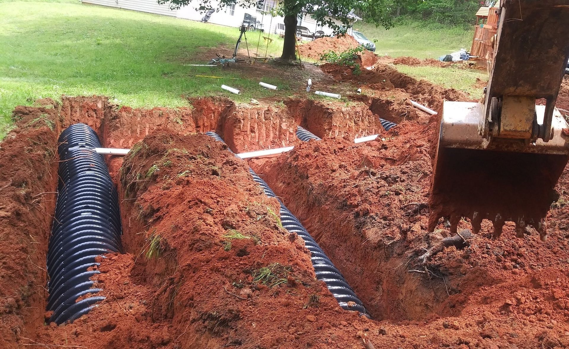 Excavator digging a trench for black drainage pipes in a muddy yard with grassy edges
