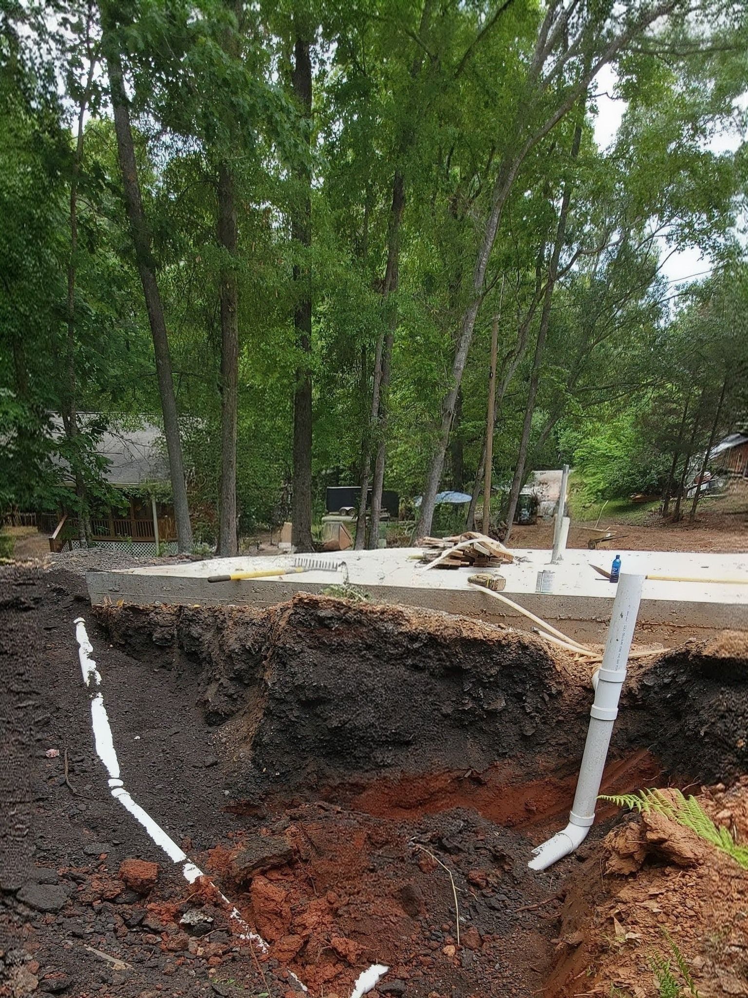 Tree-lined excavation site with exposed dark soil and white survey markers near a construction area