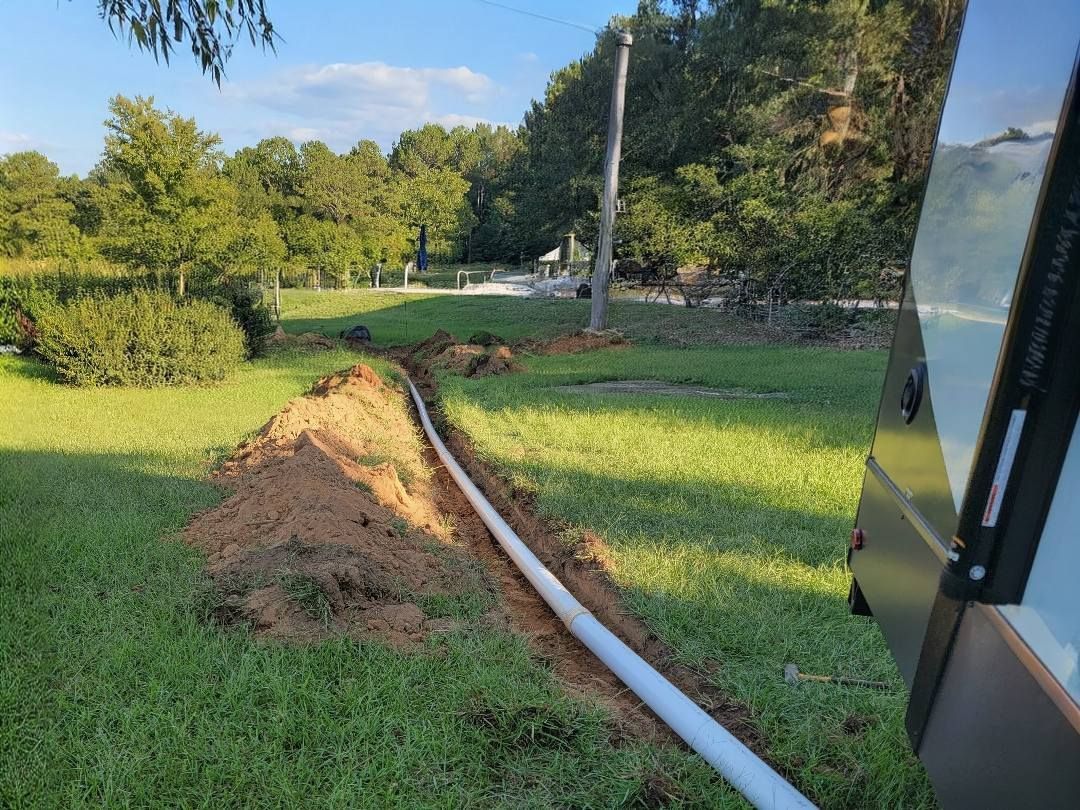 White pipe laid in a grassy yard beside a dirt trench, with trees and a building edge at right.