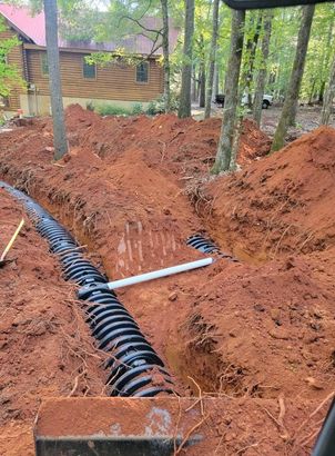 Open trench with black drainage pipe and red dirt beside a wooden cabin in a wooded area