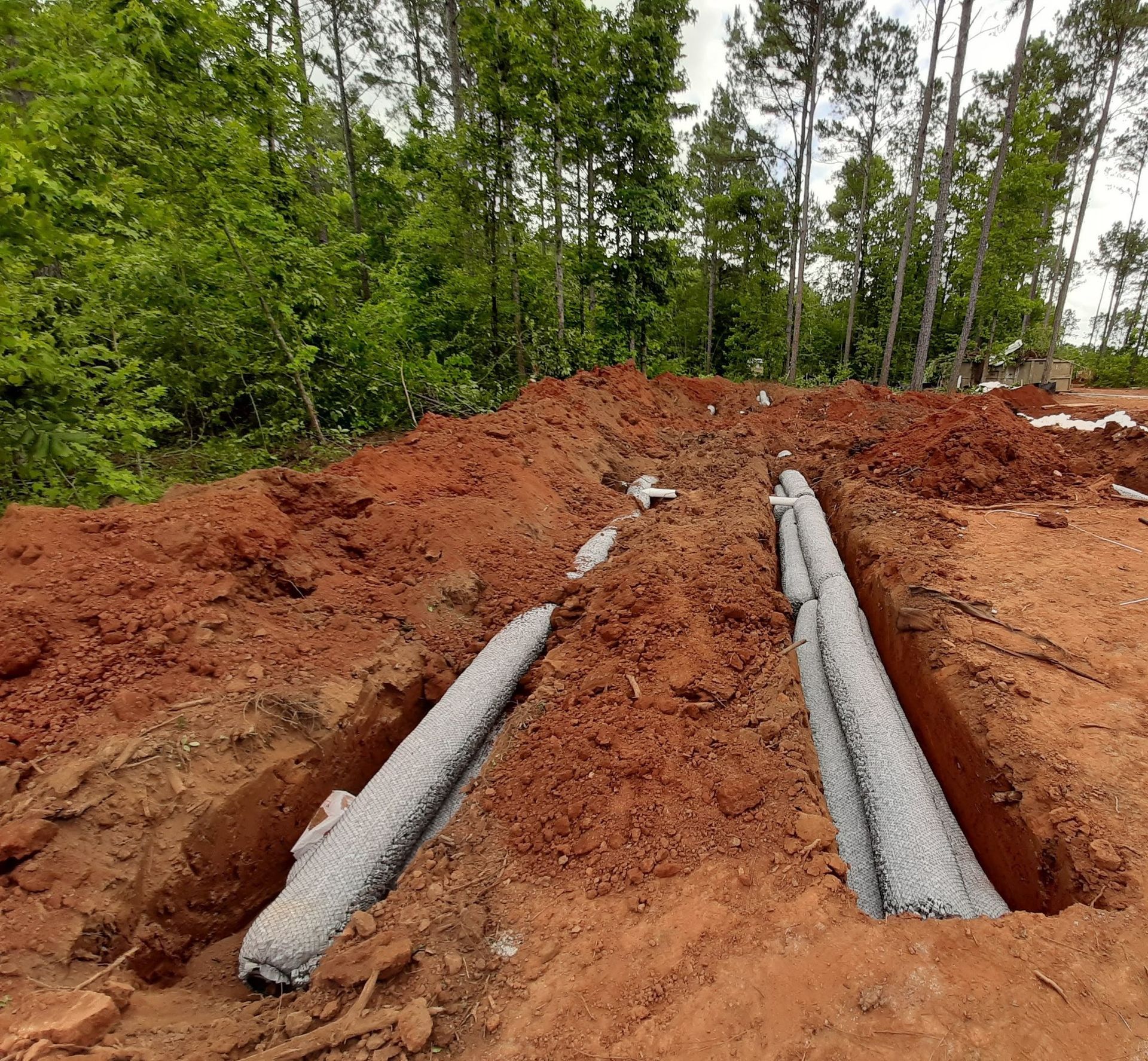 Two gray pipes laid in red dirt trench at a wooded construction site