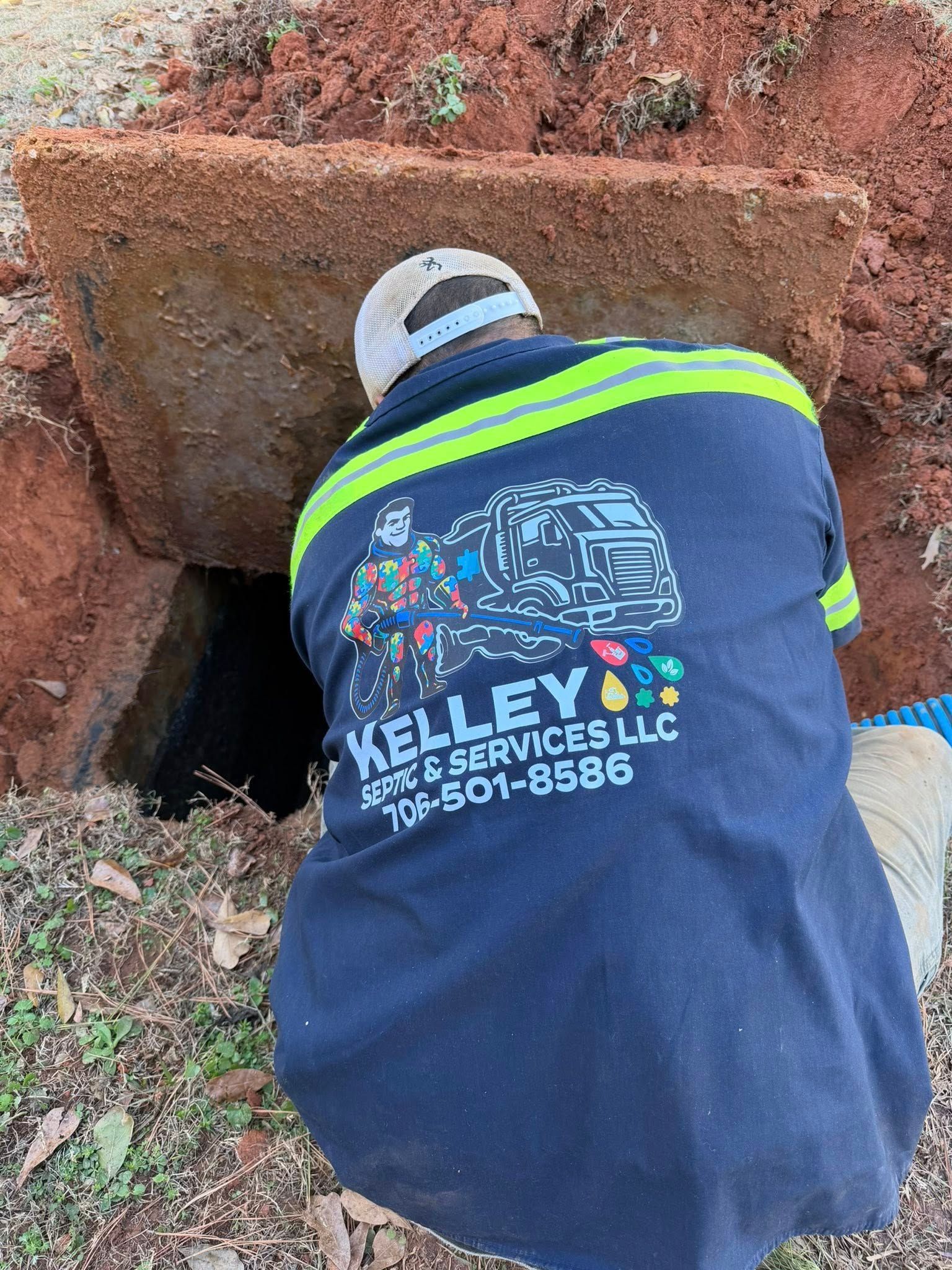 Worker in navy uniform with neon stripes kneels by a concrete drain opening outdoors.