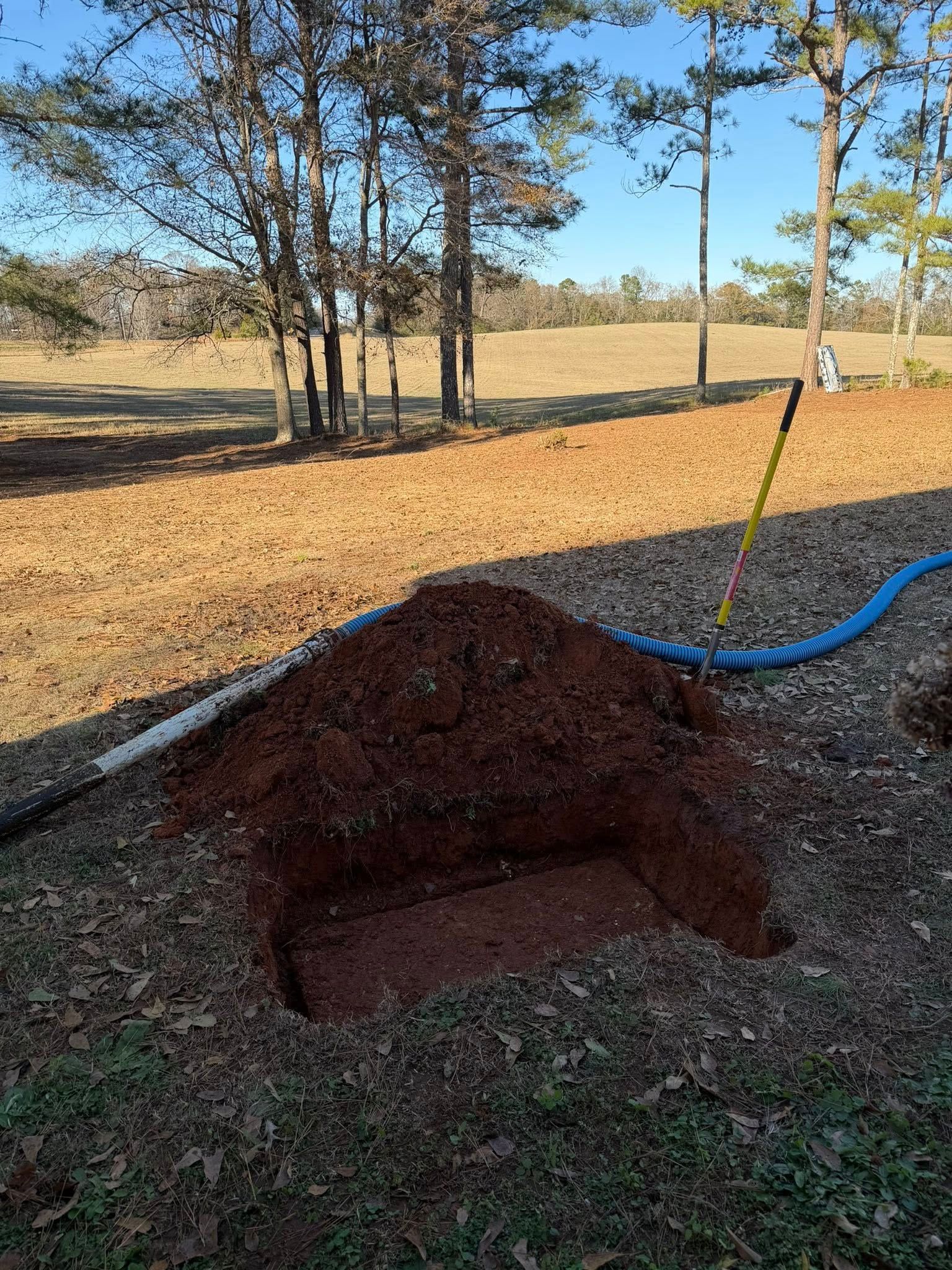 Partially dug hole in a grassy field, with a blue hose and trees in the background.