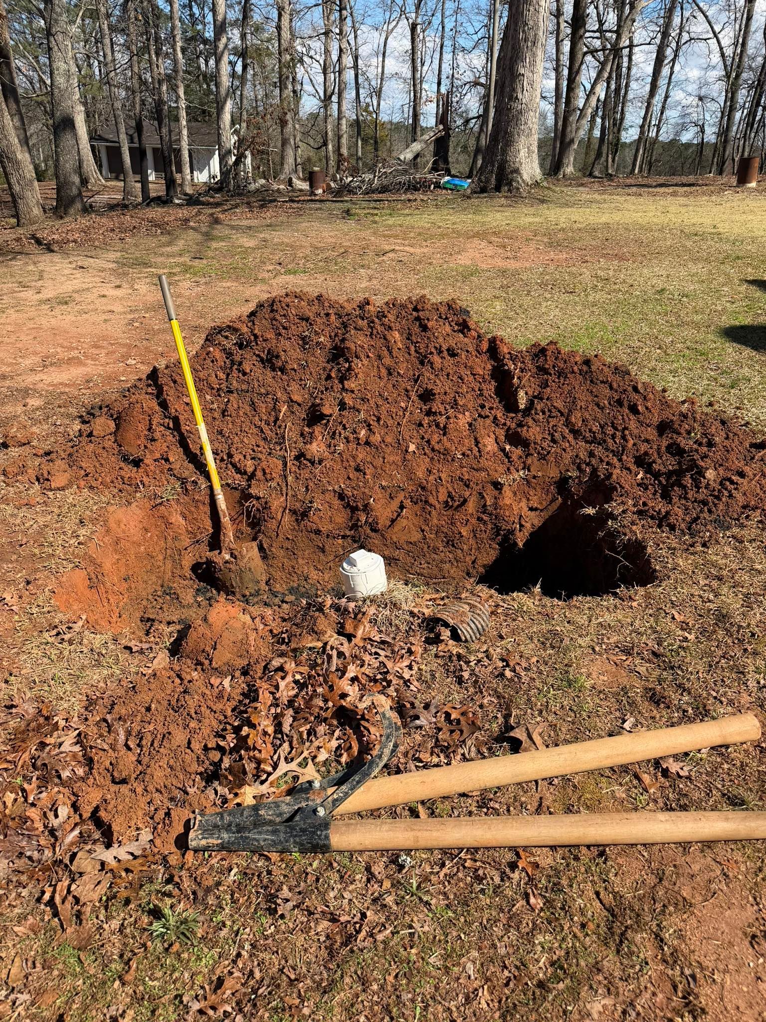 Freshly dug mound of soil beside a hole, with a shovel and rake in a wooded yard area.