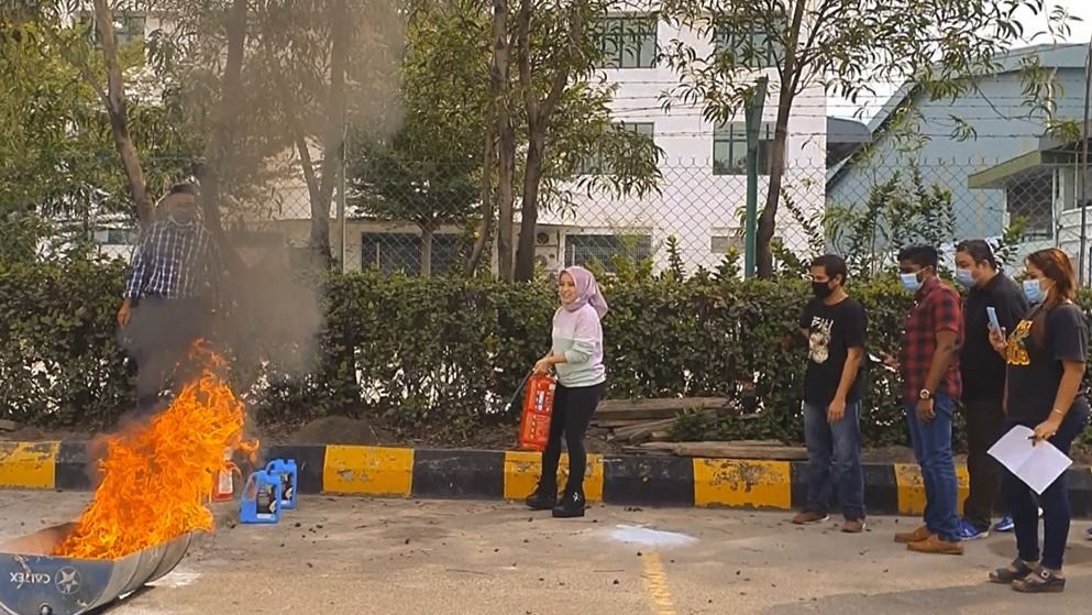 Fire extinguisher demonstration outdoors; a person using an extinguisher on a small fire, with onlookers wearing masks.