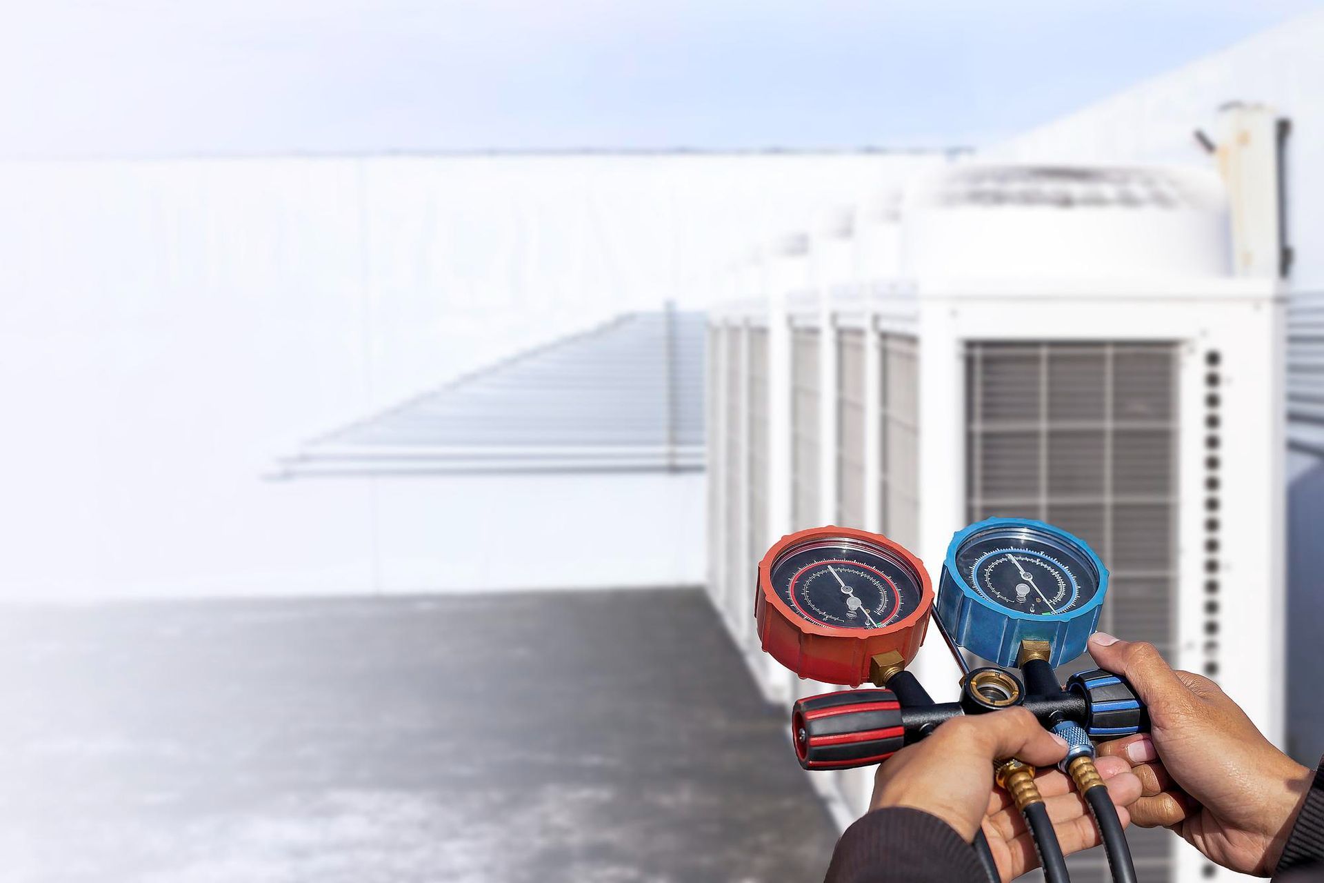 Hands holding red and blue HVAC manifold gauges near rooftop air-conditioning units