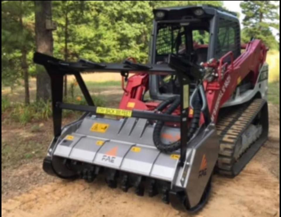 A red skid steer with a gray FAE mulching head attachment parked on a dirt path in a wooded area.