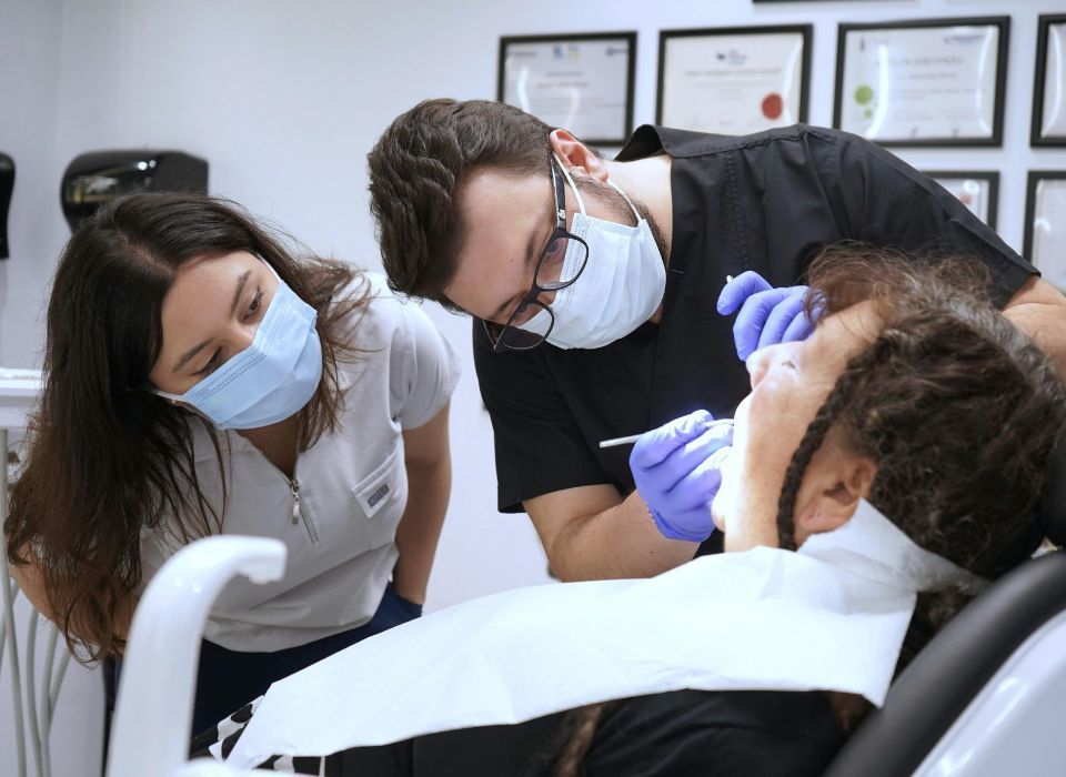 Two masked clinicians examining a patient in a dental chair under bright office lights