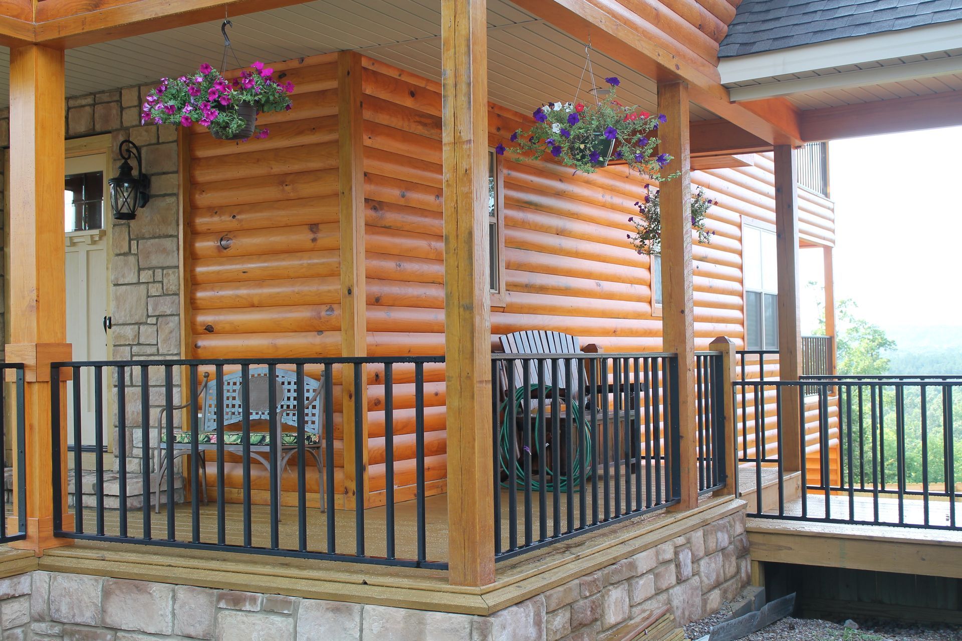 Log cabin porch with black railing, wooden beams, hanging flowers, and stone siding.