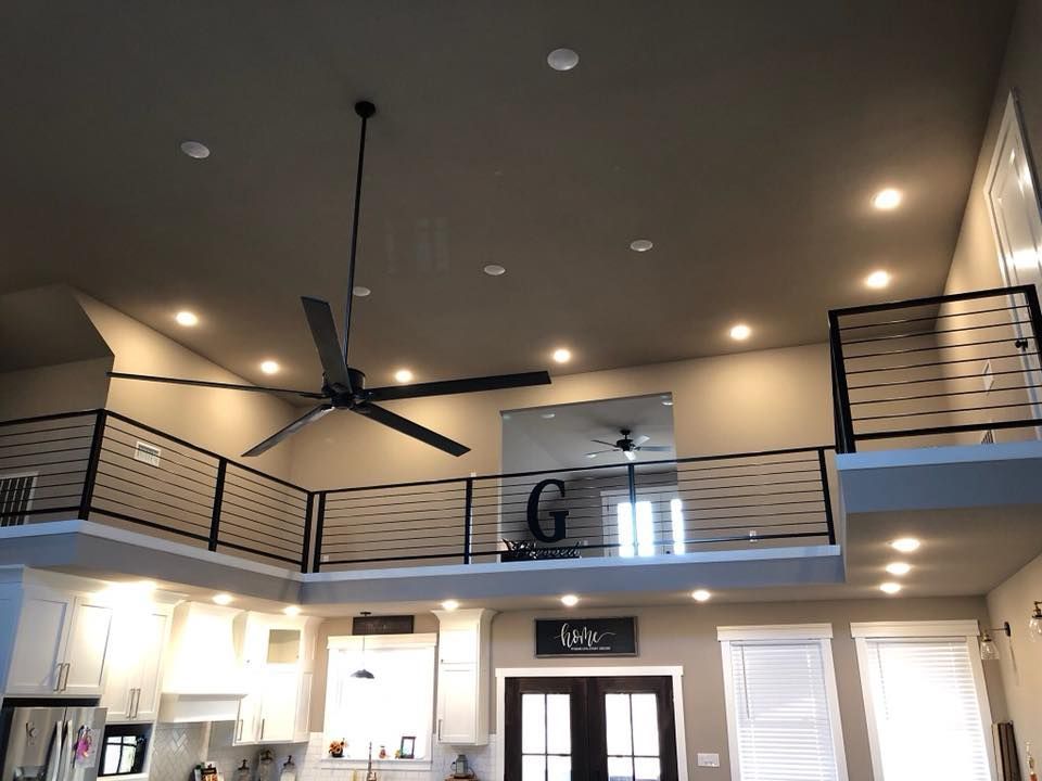 High-ceilinged home interior with a loft. Black railings, recessed lighting, and a large ceiling fan. Kitchen visible below.
