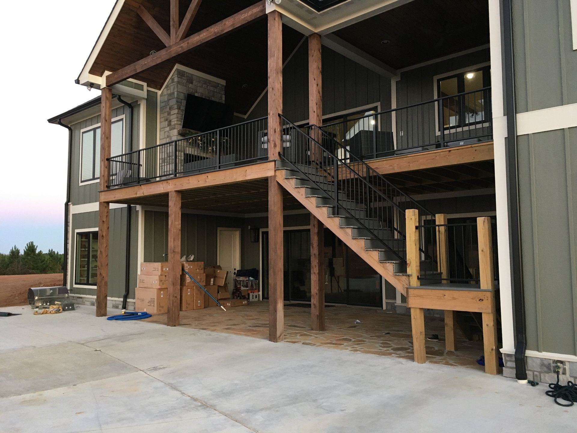 Covered outdoor patio with a wooden staircase, upper deck, and concrete driveway beside a house under construction
