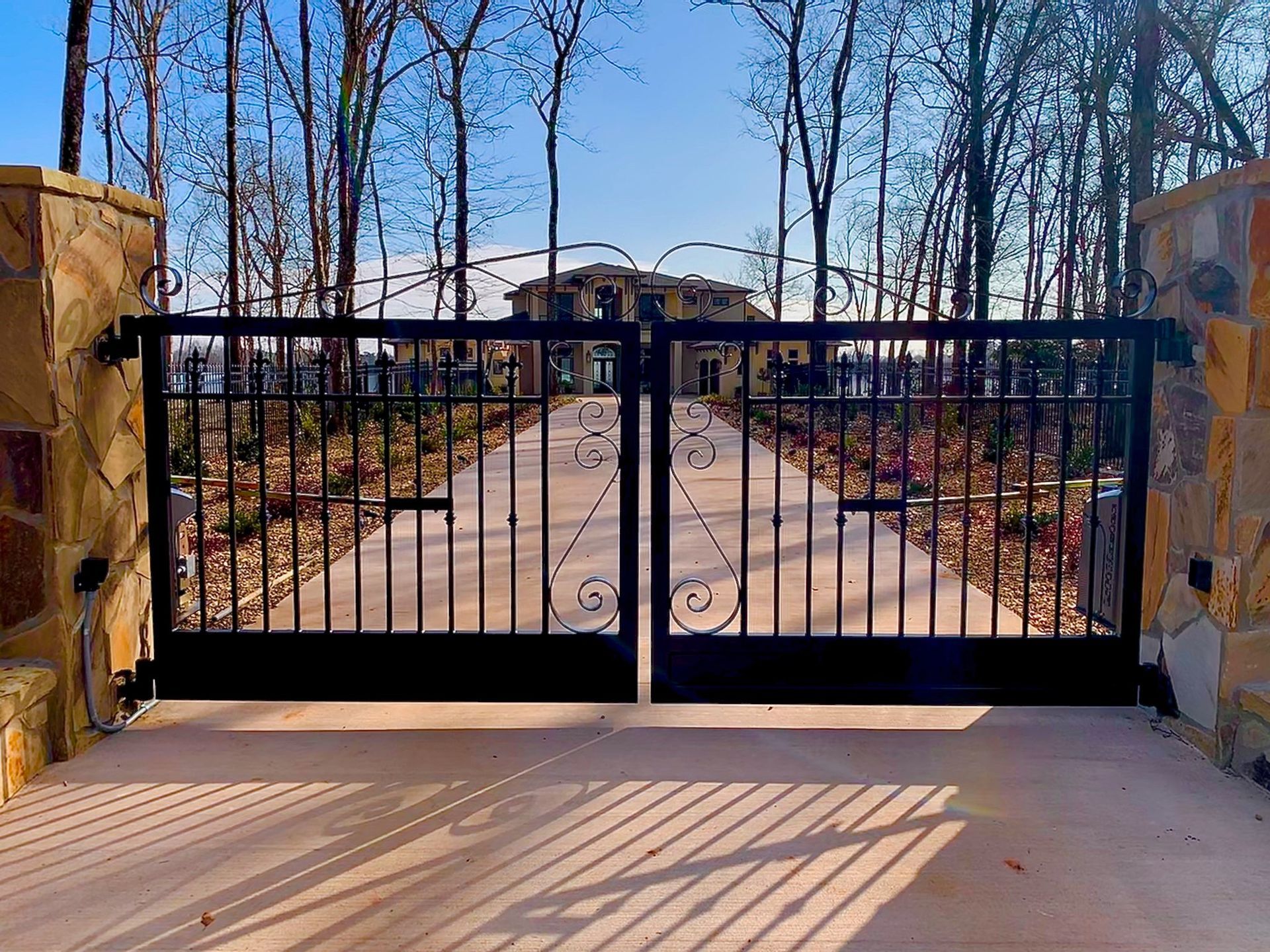 Black iron gates open to a driveway leading to a large house, flanked by stone columns and trees.