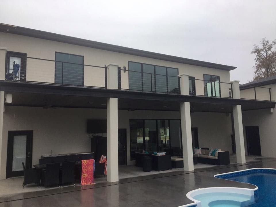 Two-story beige house with black accents. Balcony with cable railing, pool, outdoor furniture. Overcast sky.