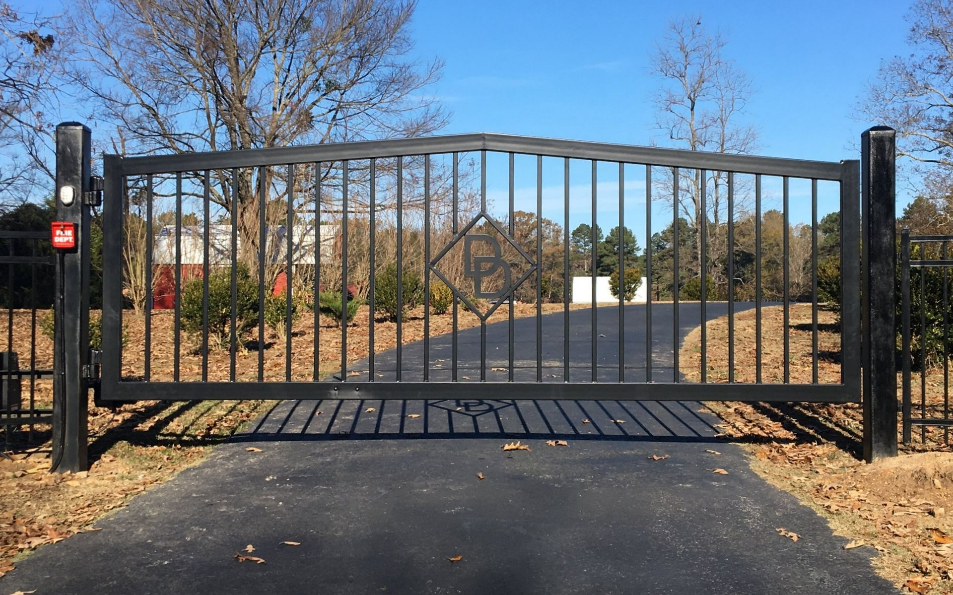 Black metal driveway gate with vertical bars and a diamond design, set on a paved road, in front of trees.