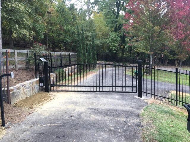 Black metal gate across a driveway, flanked by matching fencing. Trees in background.