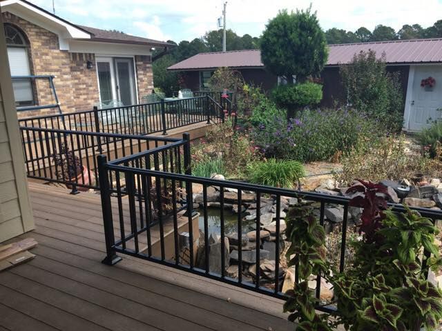 Black railing surrounds a deck with a water feature and garden in a backyard. A brick house is in the background.