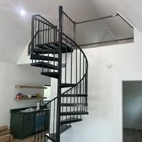 Black metal spiral staircase in a white-walled room with a small shelf and cabinets below.