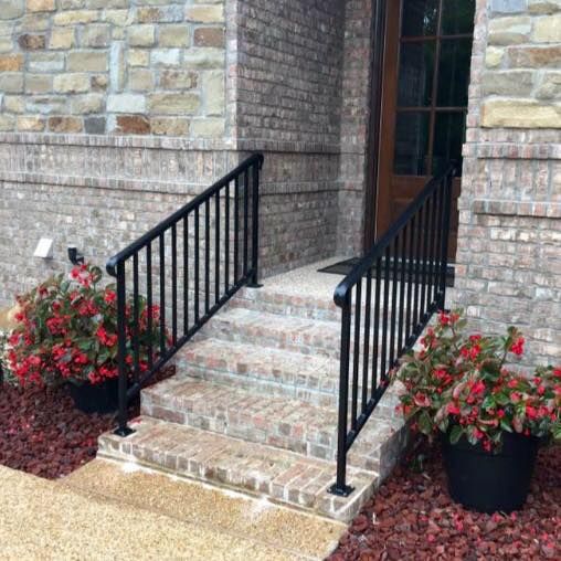 Black metal handrails flank brick steps leading to a wooden door. Red flowers in pots sit on either side.