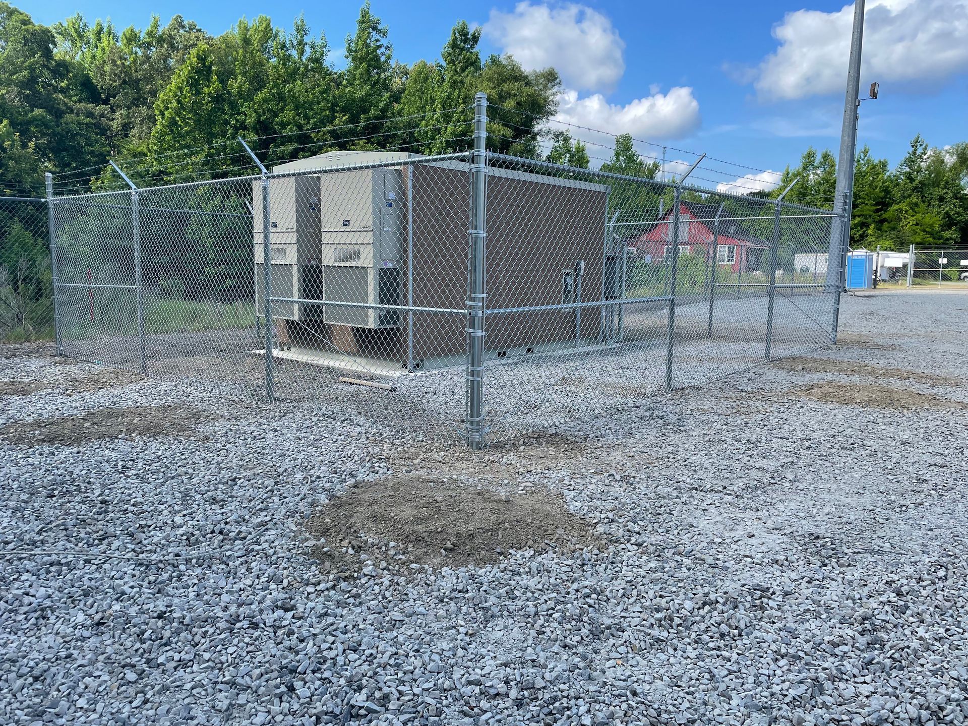 A fenced-in, gray utility box on a gravel lot; trees in the background under a blue sky.