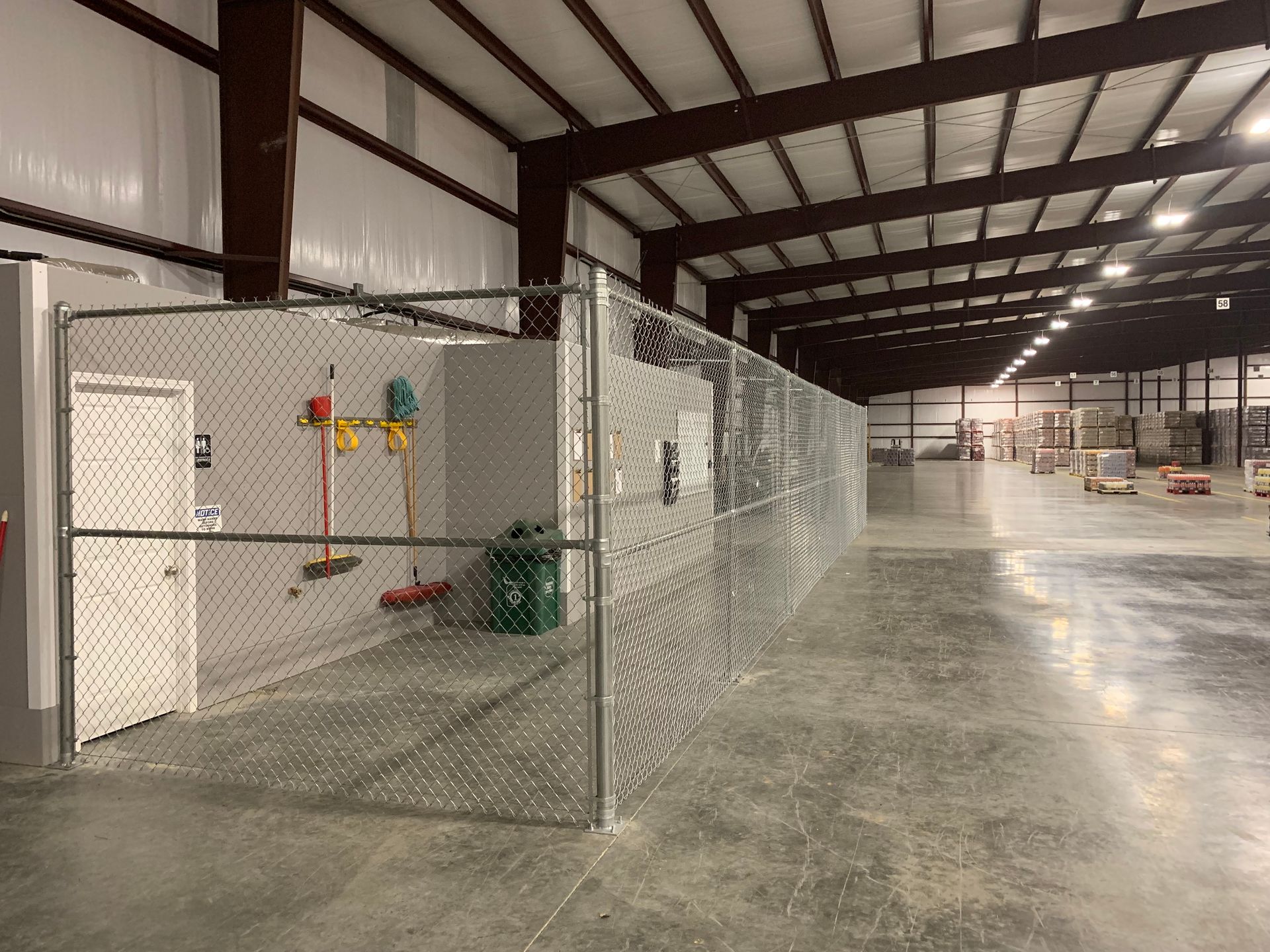 Interior of a warehouse with a chain-link fenced-in area containing a door, tools, and a green trash bag.