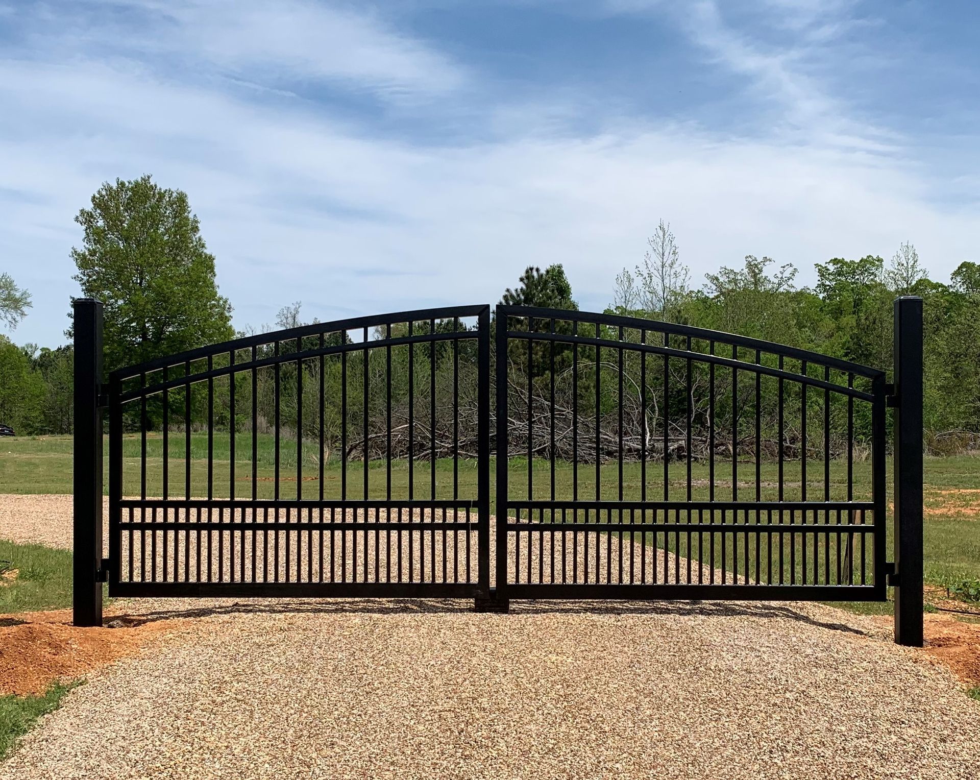 Black metal driveway gates, arched top, gravel path, open to rural scene.