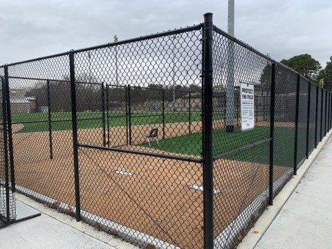 Black chain-link fence surrounds a small baseball field with brown dirt infield and green turf outfield.