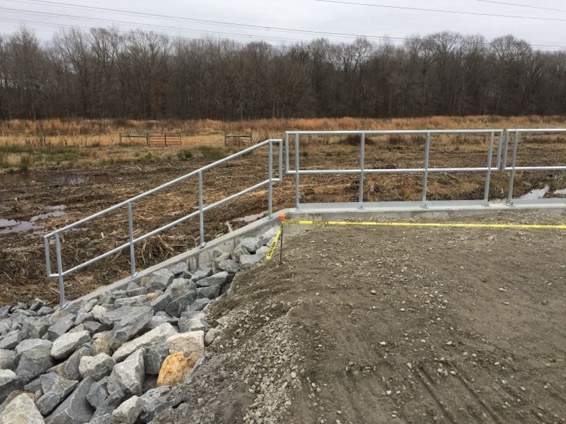 Metal railing on a retaining wall with rocks and dirt. Trees in the background. Cloudy day.
