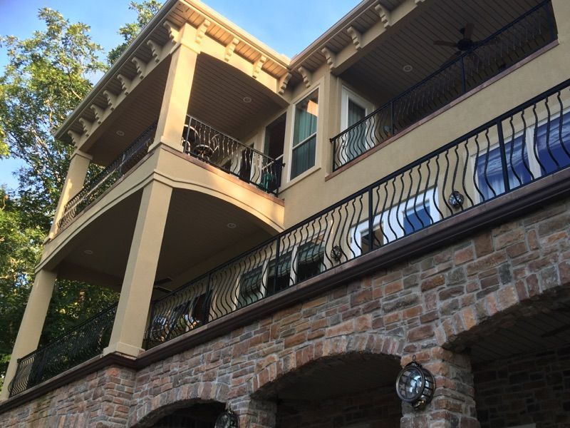 Two-story beige house with stone base, balconies, and black wrought iron railings.