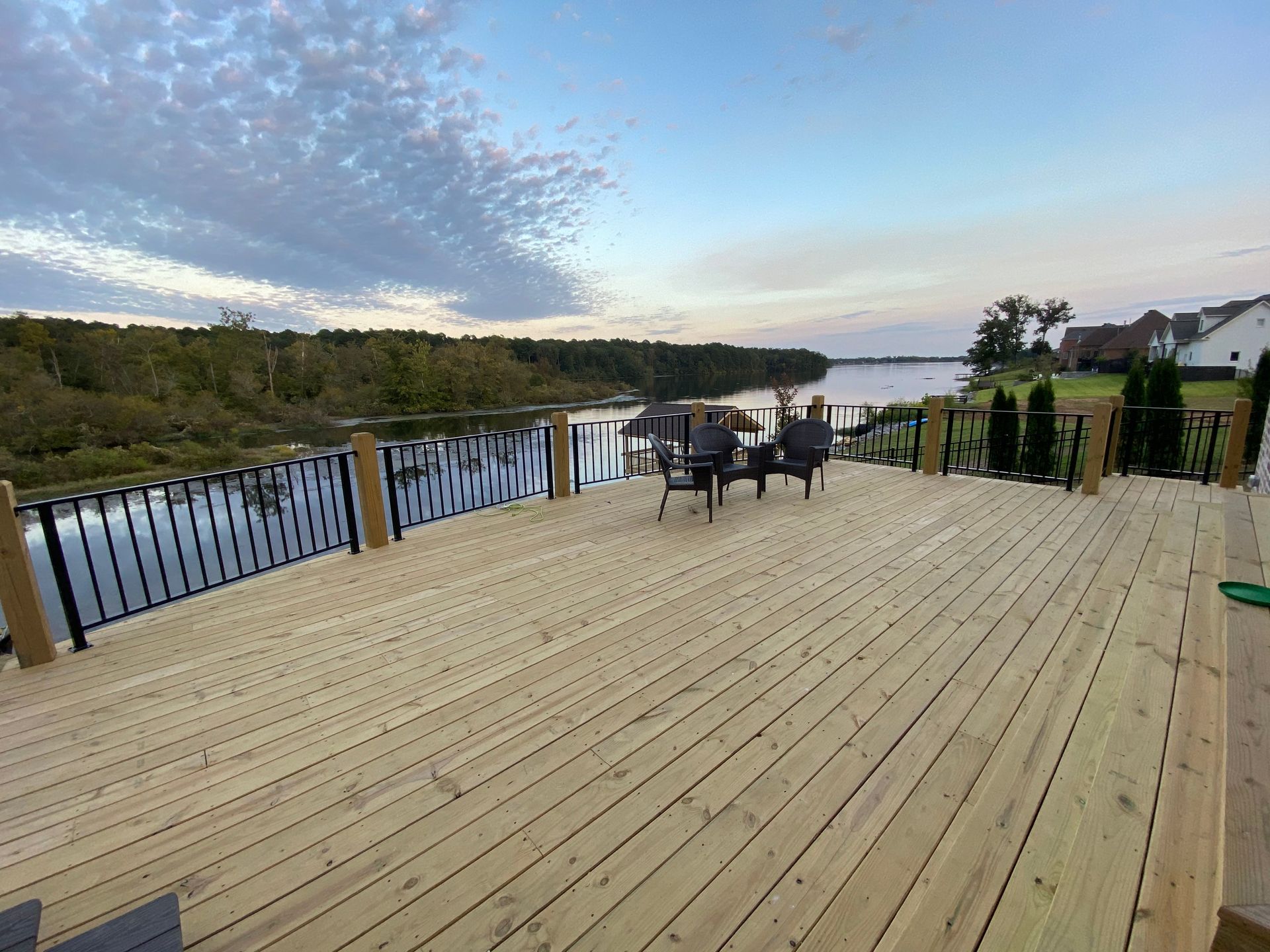 Wooden deck overlooking a calm river, trees in the distance, blue sky with clouds. Two chairs on the deck.