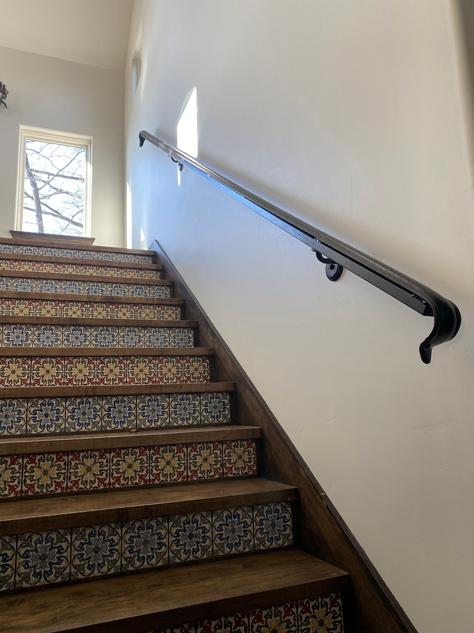 Staircase with patterned tile risers, wooden treads, and black handrail on white wall.