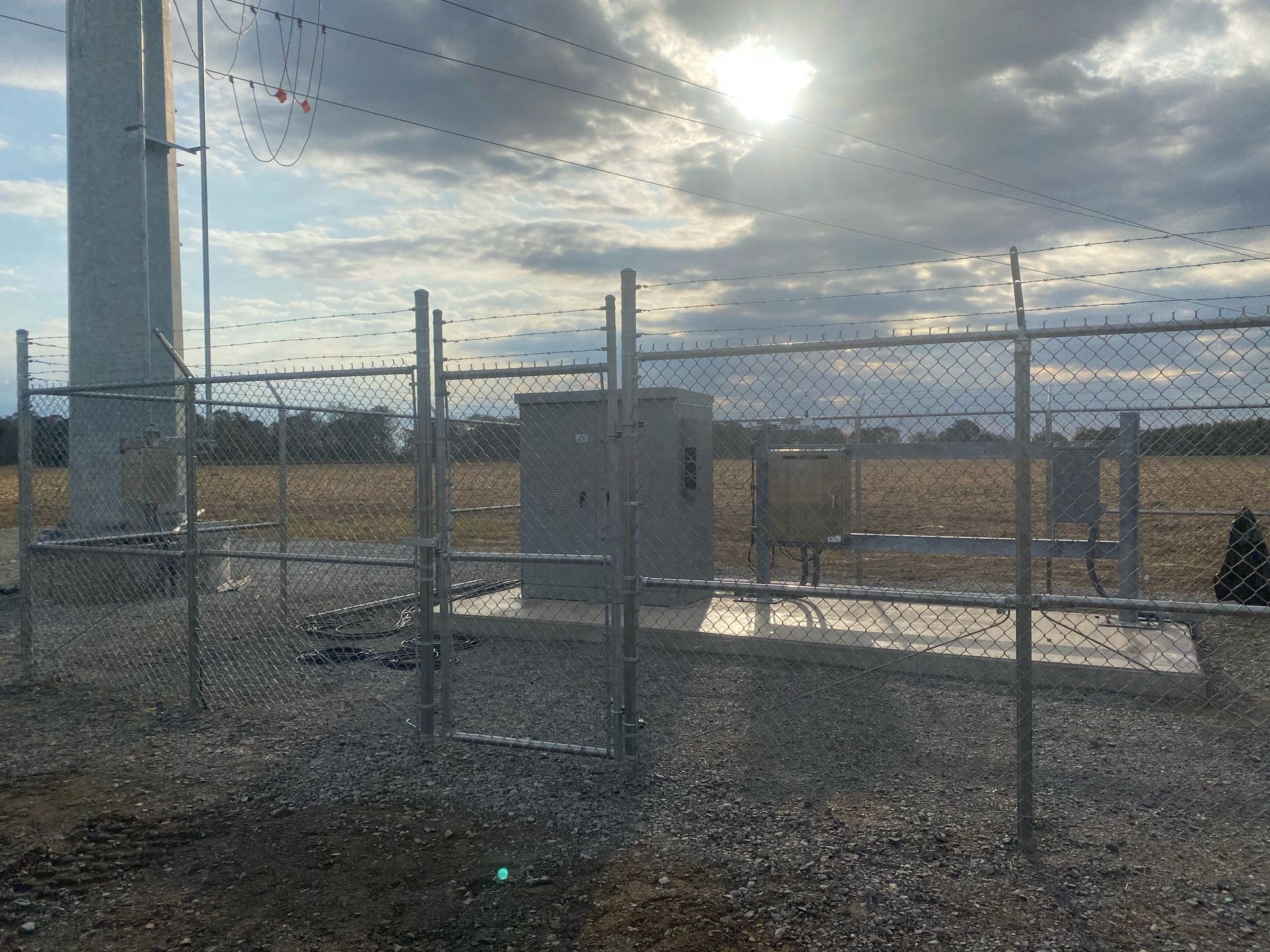 Fenced enclosure with electrical equipment next to a communication tower under a cloudy sky.