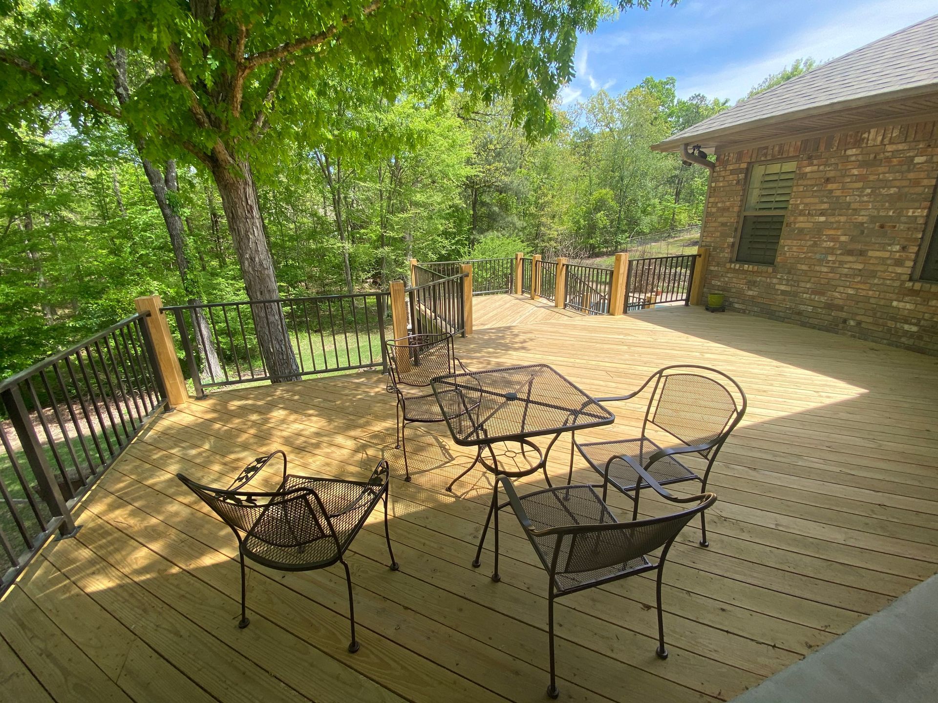Outdoor deck with metal patio chairs and table beside a brick house, surrounded by trees and a railing