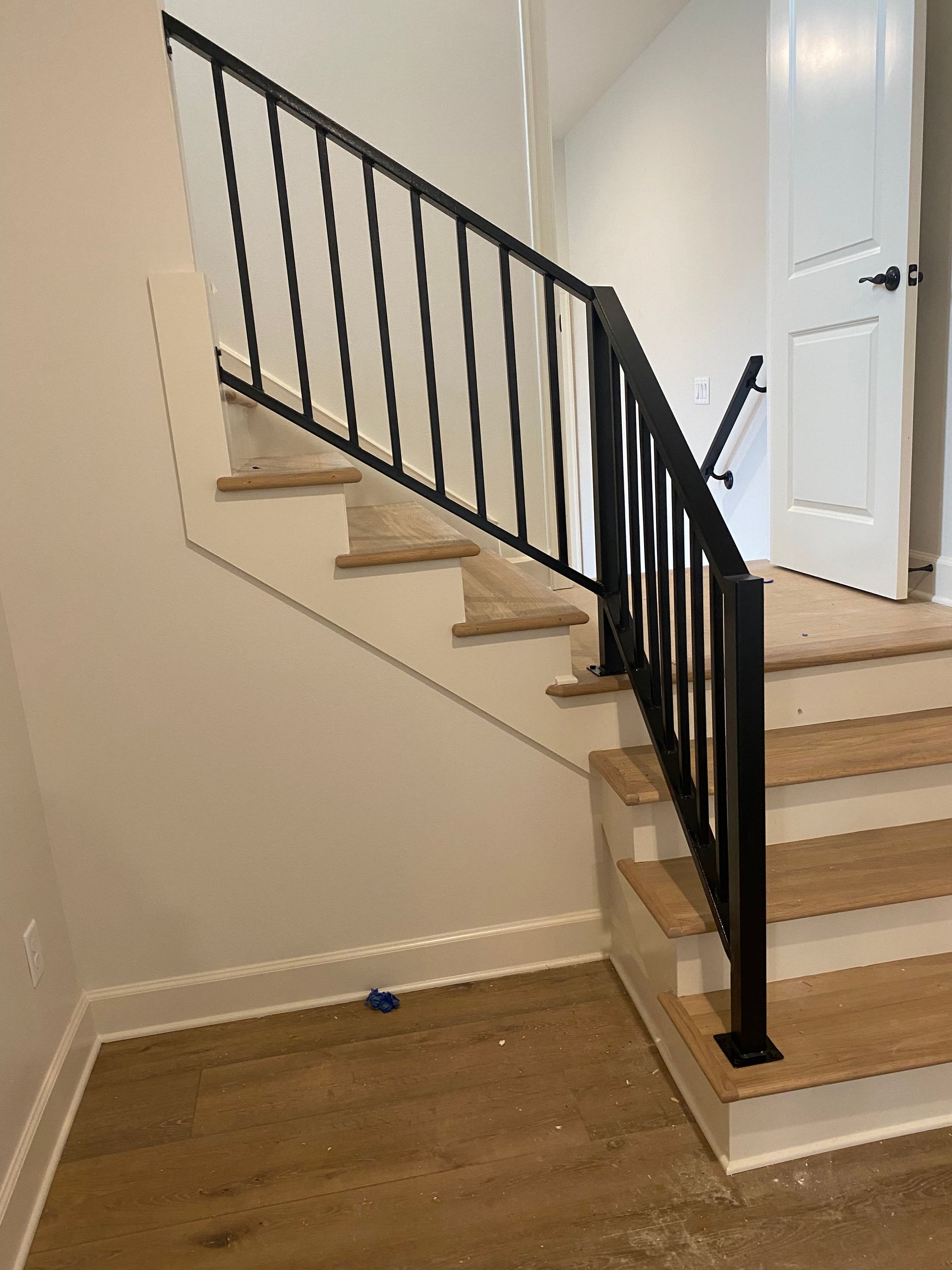 Staircase with black metal railing. Wooden steps with light brown flooring. White wall and door.