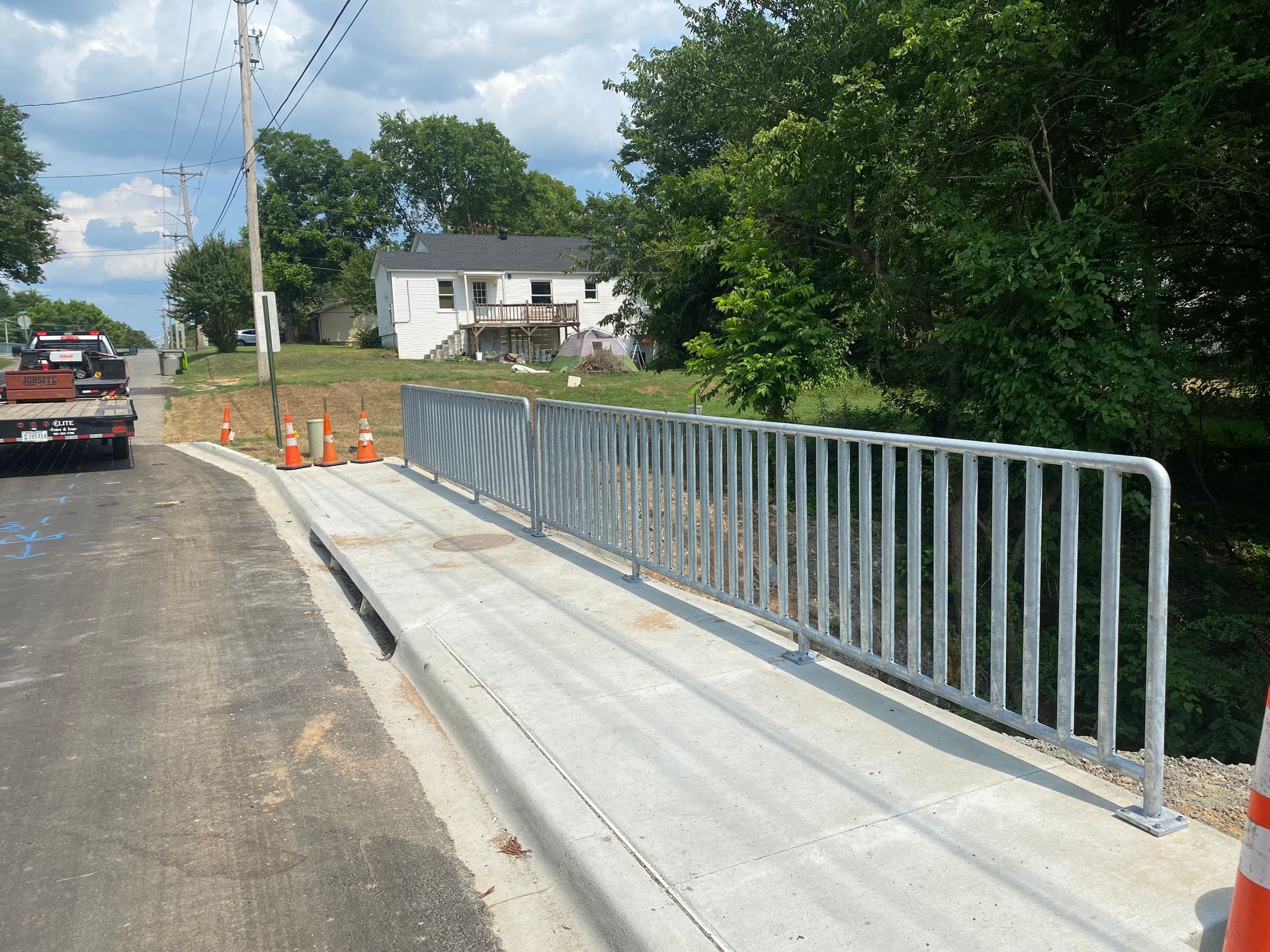 Metal railing on a concrete bridge next to a road, with a house and trees in the background.