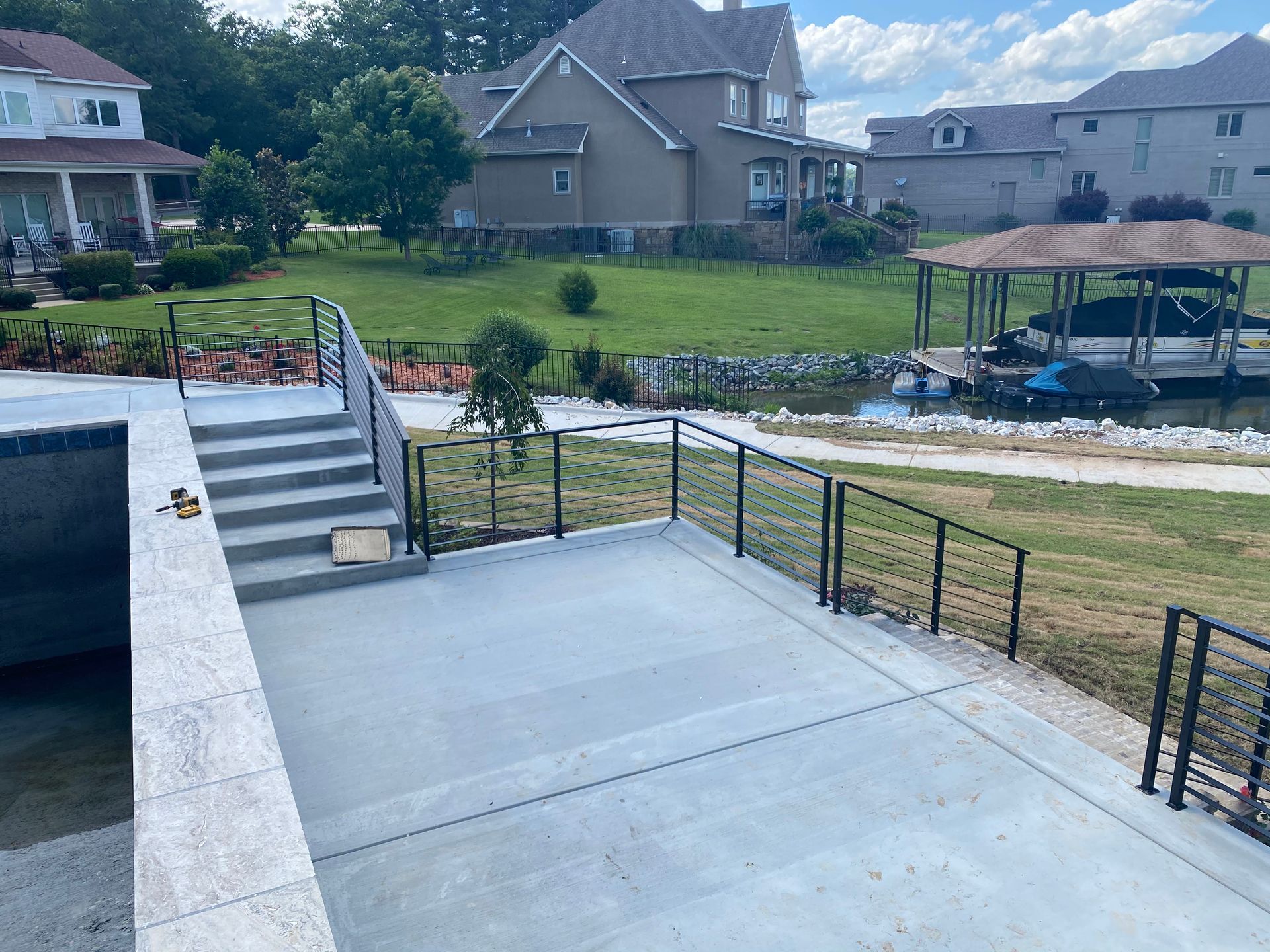 Concrete patio overlooking grassy yard with stairs, black railings, and canal-side homes in the background