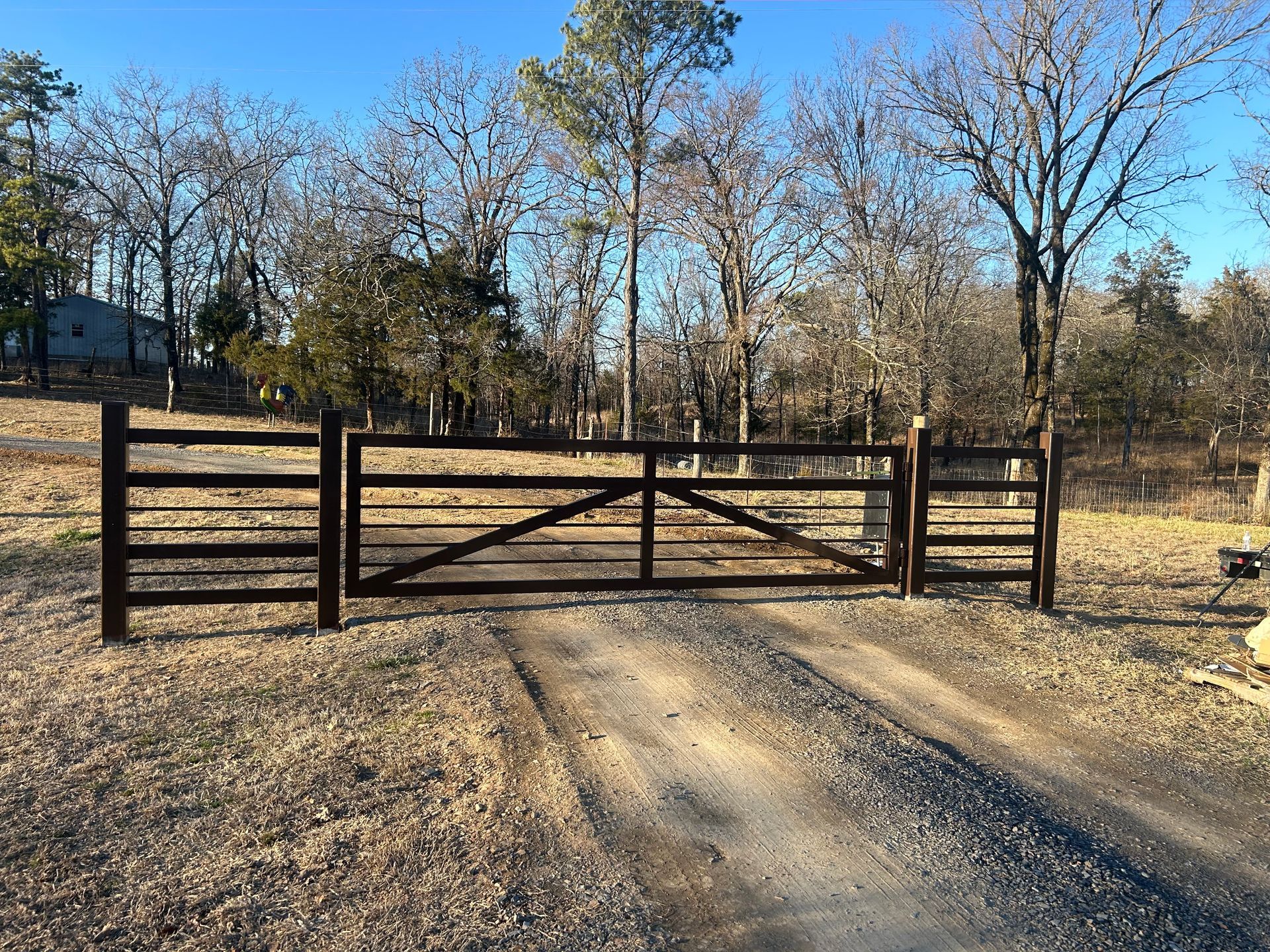 Brown wooden gate across a gravel driveway, trees in background, sunny day.