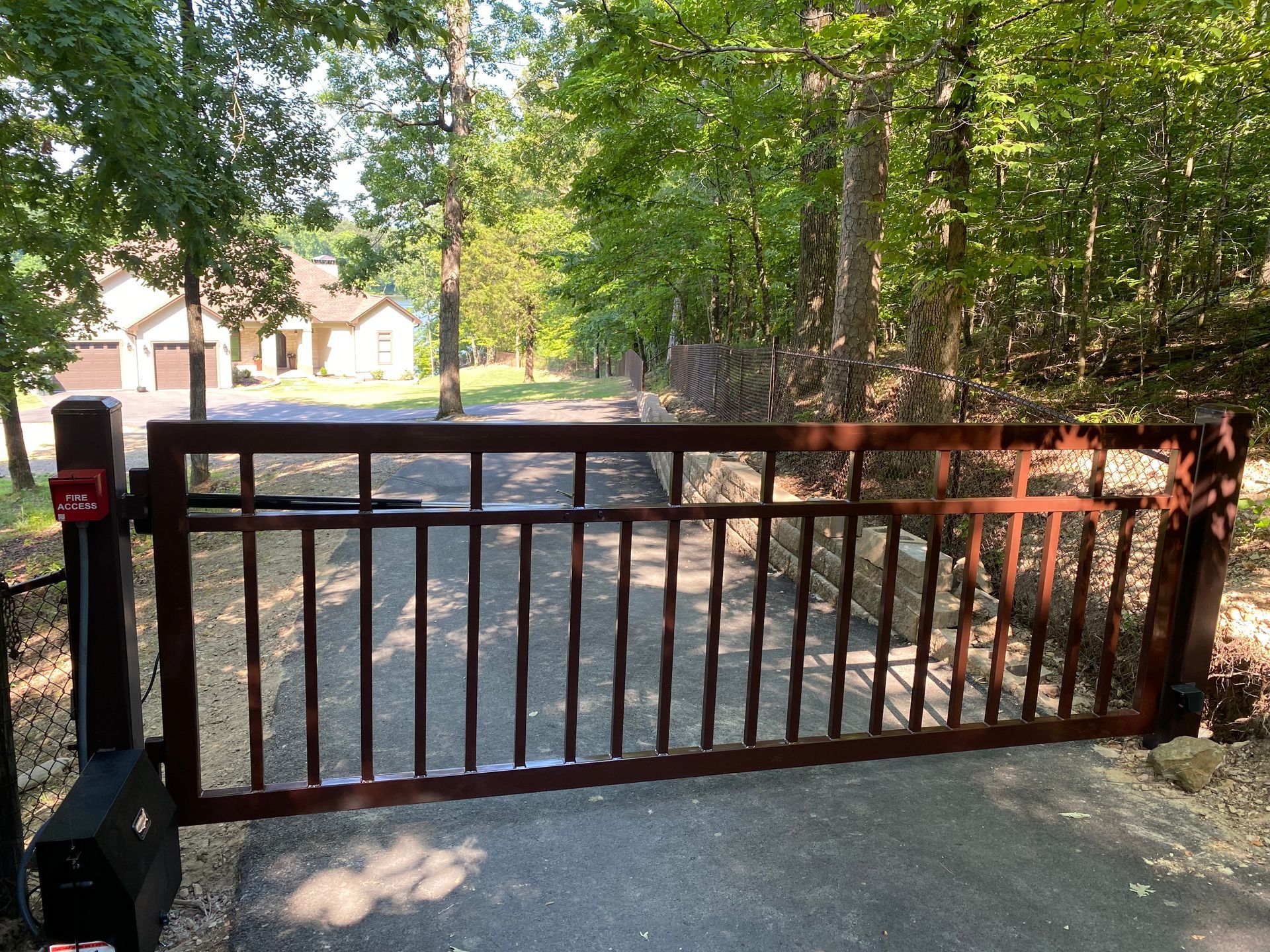 Brown metal security gate in driveway, leading to a house. Trees surround the road.
