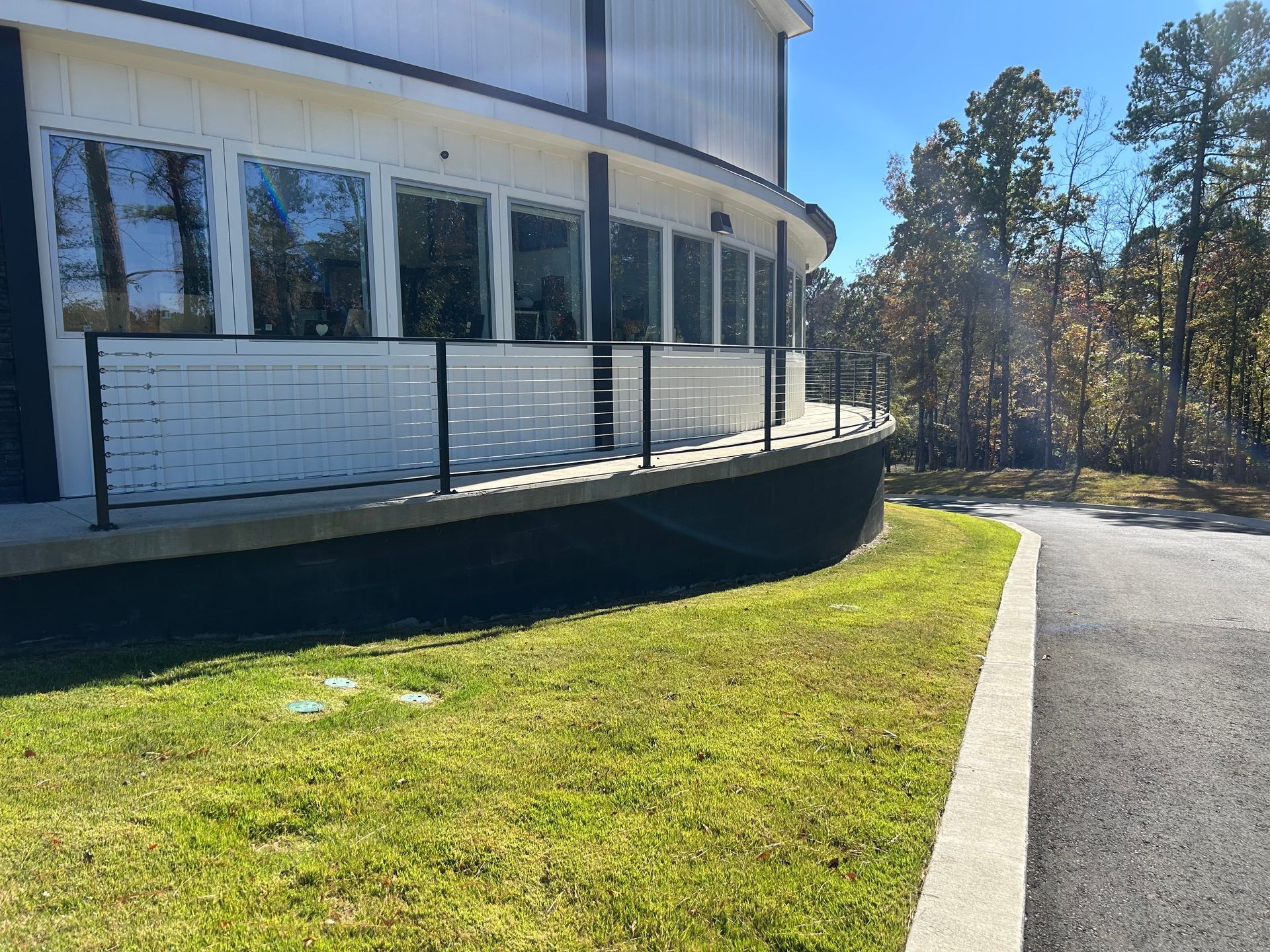Curved white building with windows, black base, and a grassy lawn beside a paved path on a sunny day