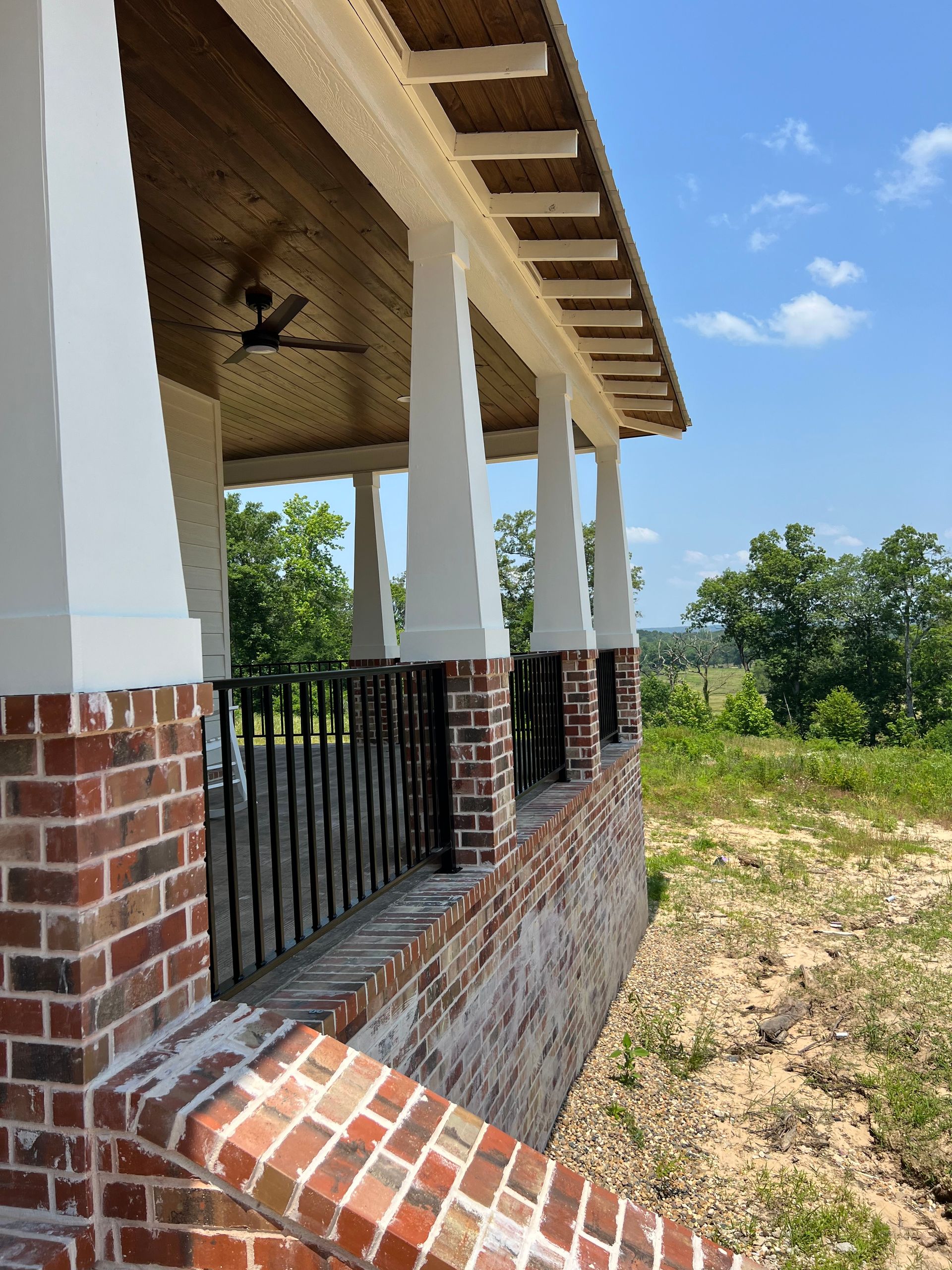Brick porch with white columns, black railing, and a view of green trees under a blue sky.