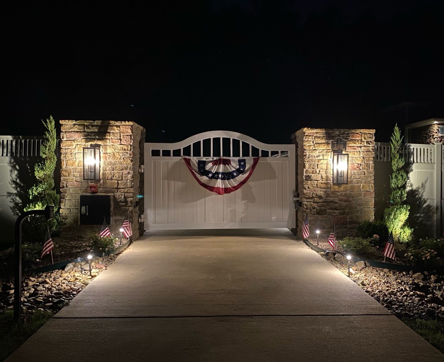 A white gate decorated with a patriotic banner is lit up at night. The gate is flanked by stone pillars with lights.