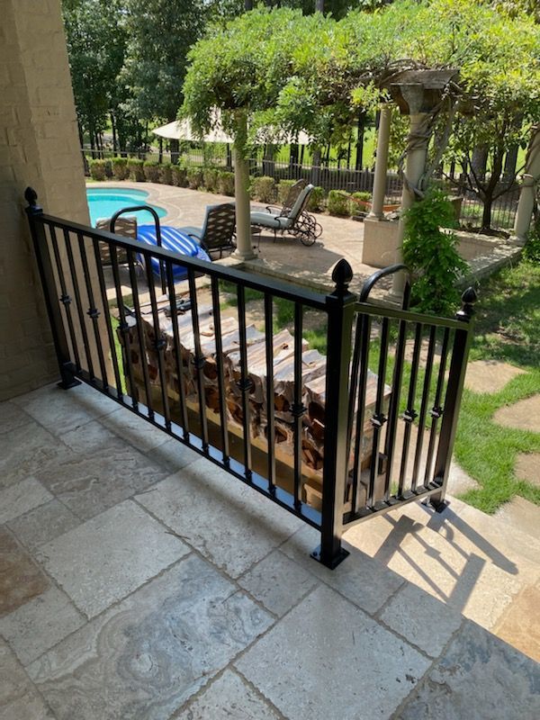 Balcony view of a backyard patio with black railing, blue chairs, and a pergola among trees and lawn