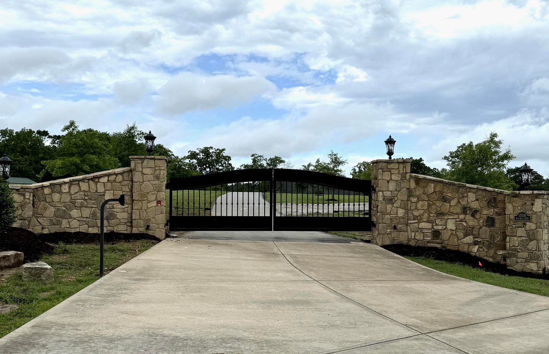 Stone pillars with black wrought iron gate on a concrete driveway under cloudy sky.