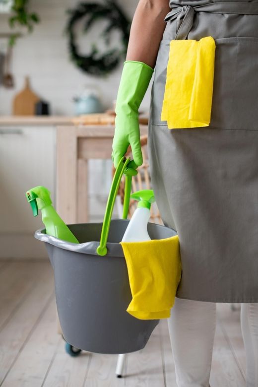 Person smiling in a bathroom, holding cleaning supplies and a cloth near the sink and toilet.