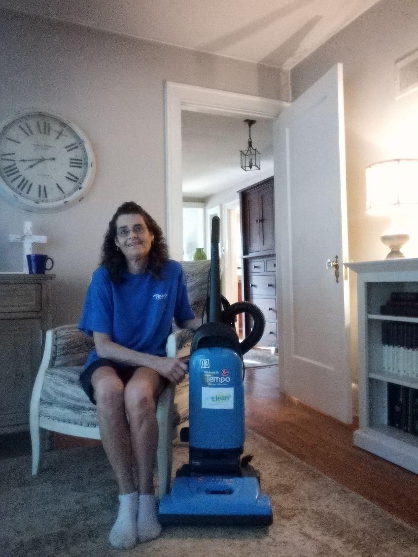 Woman sits beside a blue vacuum cleaner in a living room with fireplace and wall clock.