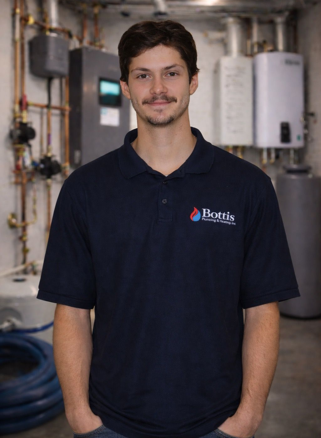 Man standing in a workshop, wearing a navy polo shirt with hands in pockets.