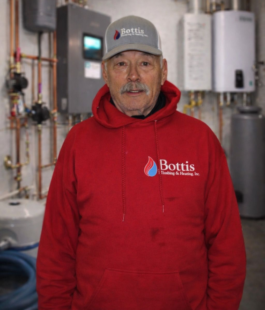 Person in a red Bottis hoodie and gray cap standing in a mechanical room with pipes and water heaters.