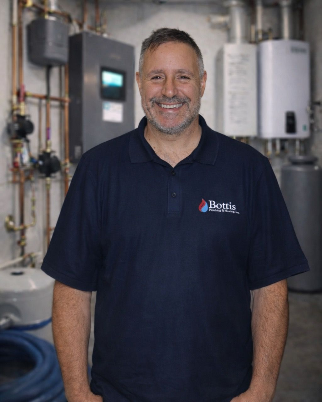 Smiling technician in navy polo stands in a utility room with water heaters and HVAC equipment.
