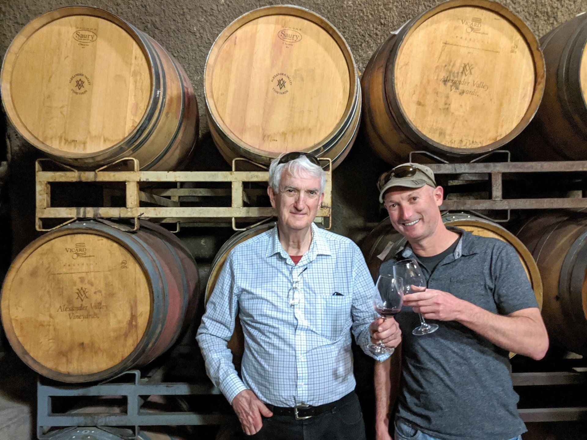 Two men are standing next to each other in front of a stack of wine barrels.