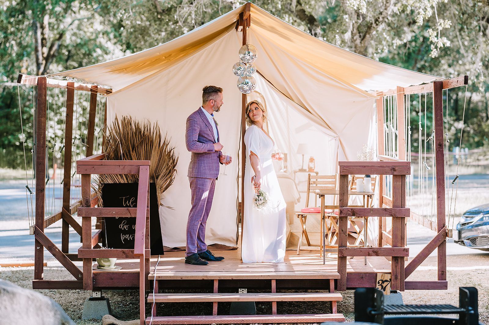Man and women dressed in wedding attire standing in on the deck in front of a glamping tent